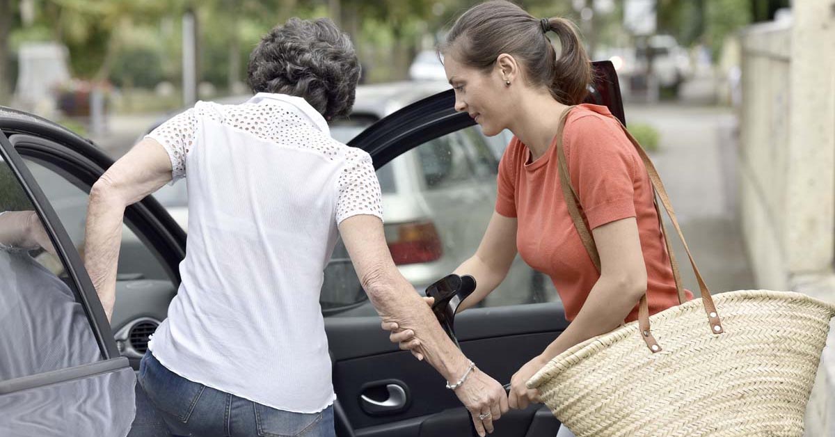 A young carer holding the hand of an older person to help them into a car.