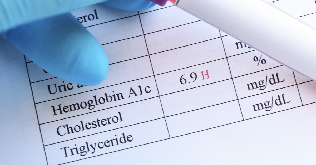 A gloved hand holding a blood sample in a test tube and hovering above a piece of paper recording HbA1c levels.