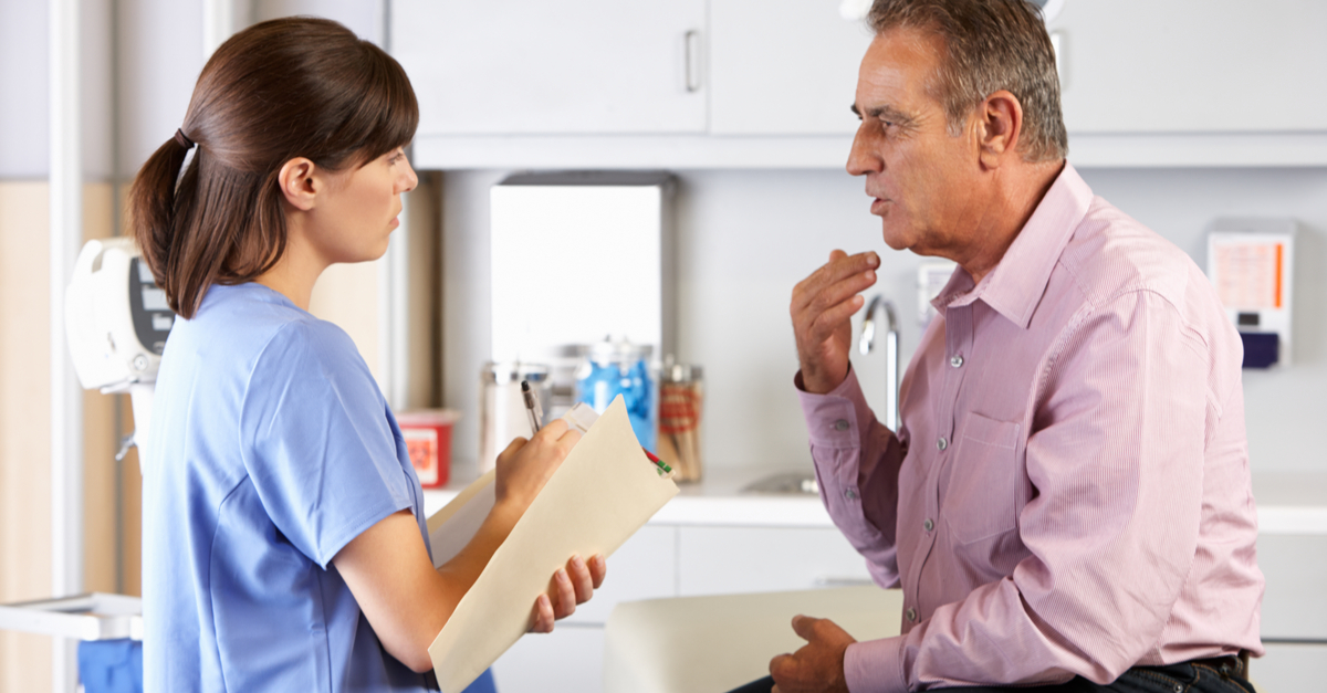 An older patient gesturing towards his face to indicate a health issue while receiving care from a doctor or nurse in a general practice.