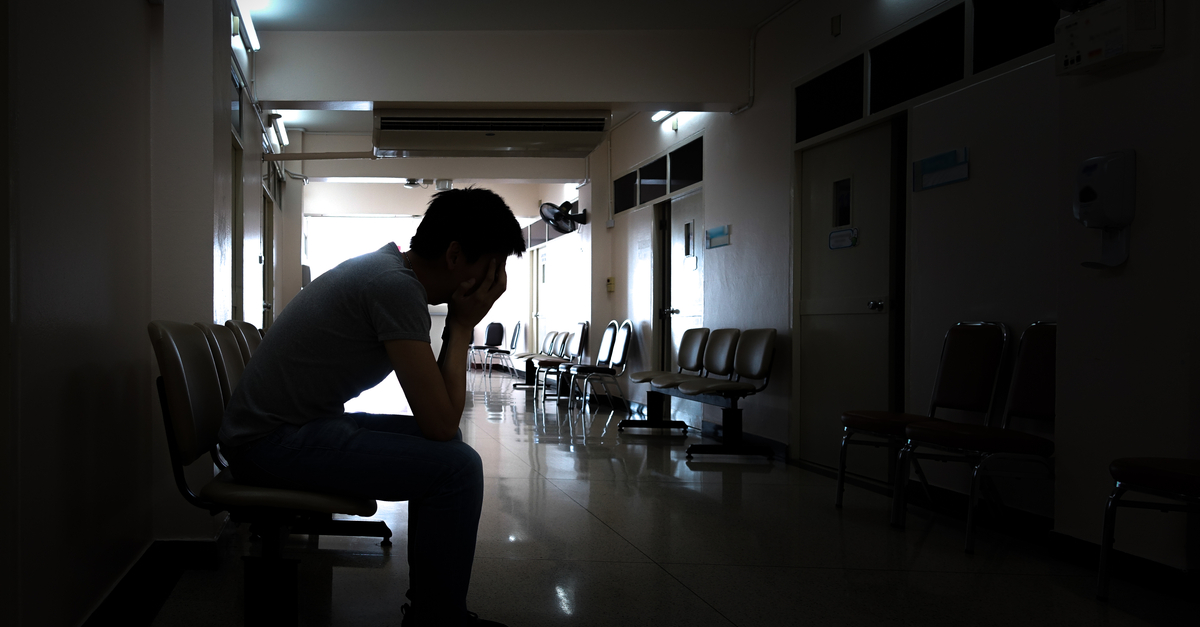 A patient sitting with their head in their hands in a dark general practice waiting room.