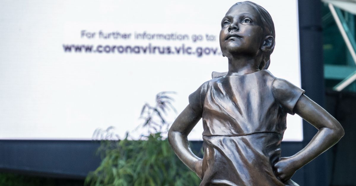 Photograph of statue of girl striking defiant pose in front of billboard with URL www.coronavirus.vic.gov.au