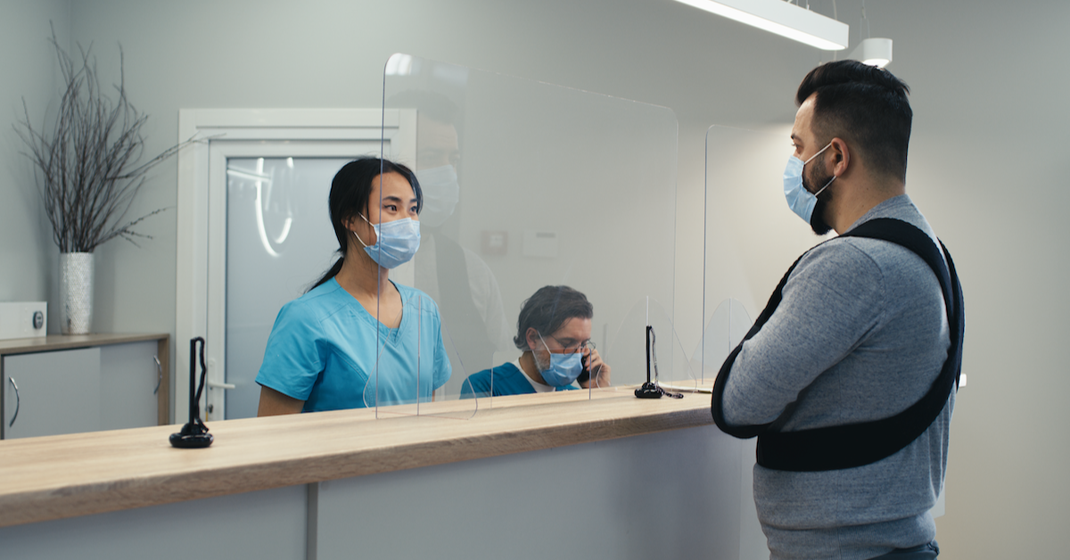 A patient with their arm in a sling speaks to a health service receptionist, both wearing masks.