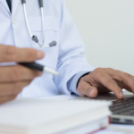 An unidentified general practitioner fills out paperwork in front of his laptop computer