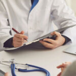 An unidentified male general practitioner writes notes as a female patient speaks with him