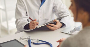An unidentified male general practitioner writes notes as a female patient speaks with him