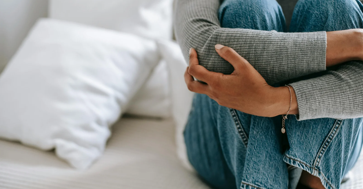 An unidentified woman sitting down hugs her knees, which are up at her chest, while sitting on a bed