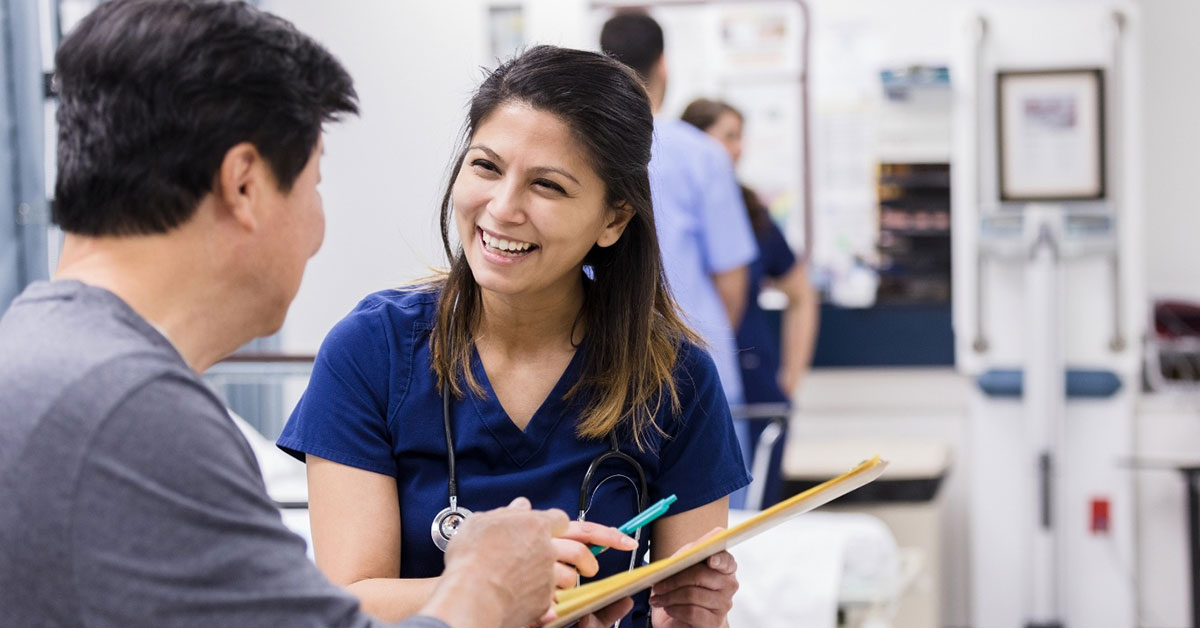 A practice nurse holds a clipboard and speaks with a middle aged Asian male patient