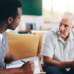 A young male nurse holding a clipboard speaks with a middle aged male patient