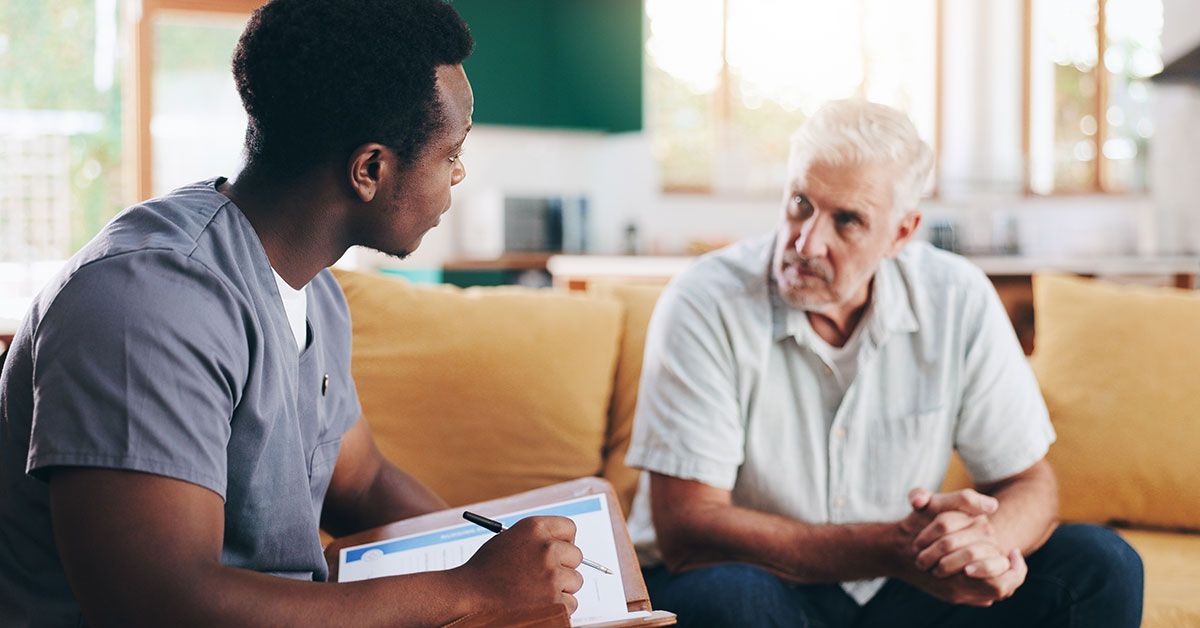 A young male nurse holding a clipboard speaks with a middle aged male patient