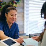 A clinician speaks with a patient filling out a form at the reception area of a GP clinic