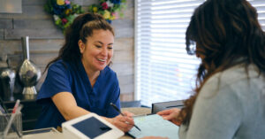 A clinician speaks with a patient filling out a form at the reception area of a GP clinic