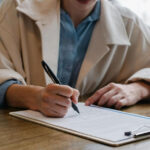 An unidentified woman sitting at a desk signs a document on a clipboard.