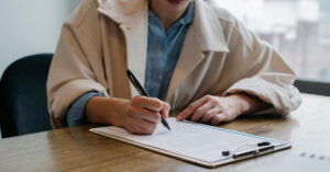 An unidentified woman sitting at a desk signs a document on a clipboard.