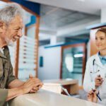 Young happy nurse taking notes while communicating with mature patient at reception desk at medical clinic.