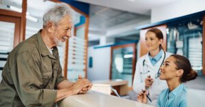 Young happy nurse taking notes while communicating with mature patient at reception desk at medical clinic.