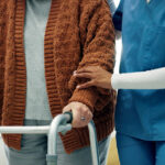 An elderly patient is assisted by a female nurse to walk with her walking frame