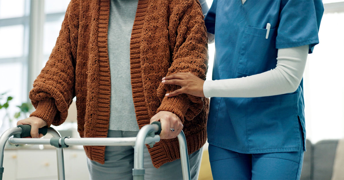 An elderly patient is assisted by a female nurse to walk with her walking frame